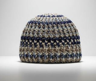 A crocheted beanie hat featuring a textured pattern of navy blue, beige, and white yarn, shown on a neutral light gray background.