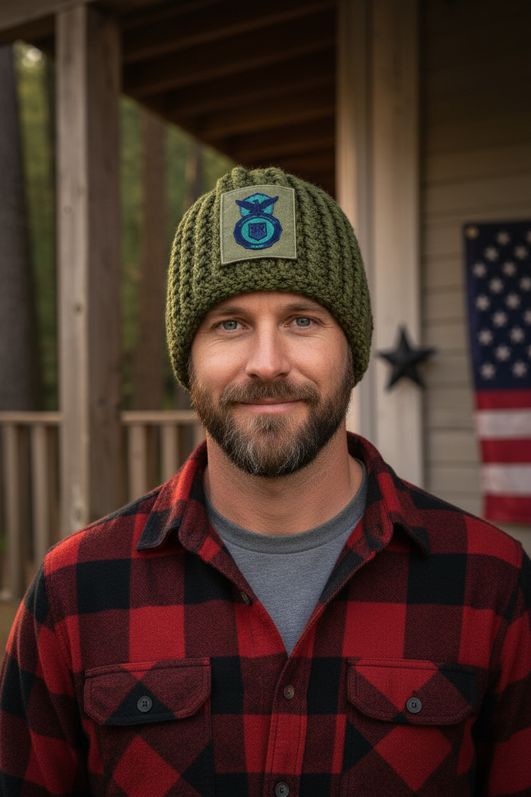 A man wearing an olive green crochet beanie with a blue and black military-style insignia patch on the front, paired with a red and black buffalo plaid flannel shirt, standing on a wooden porch with an American flag in the background.