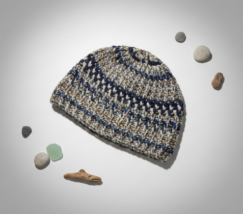 A crochet beanie hat in a variegated pattern of navy blue, beige, and cream, styled flat-lay on a light gray background with small decorative stones.