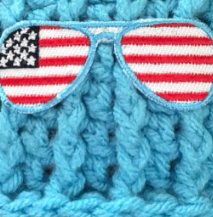 A close-up of a bright blue chunky crochet beaine featuring a decorative embroidered patch shaped like aviator sunglasses with an American flag design.