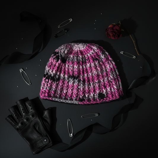 Pink and gray crochet beanie with black accents on a dark surface with gloves and a rose.