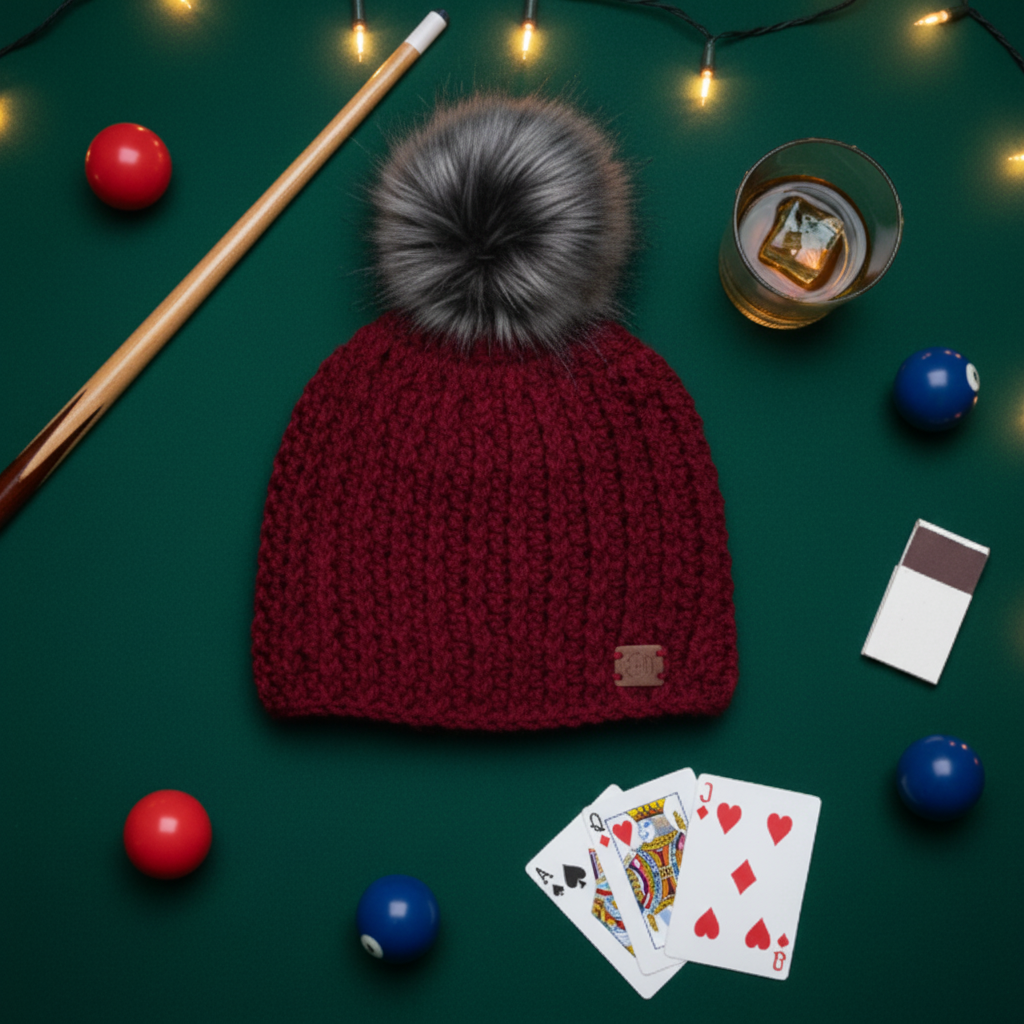 Red crochet beanie with gray pom-pom on a green surface with pool cues, balls, and playing cards.