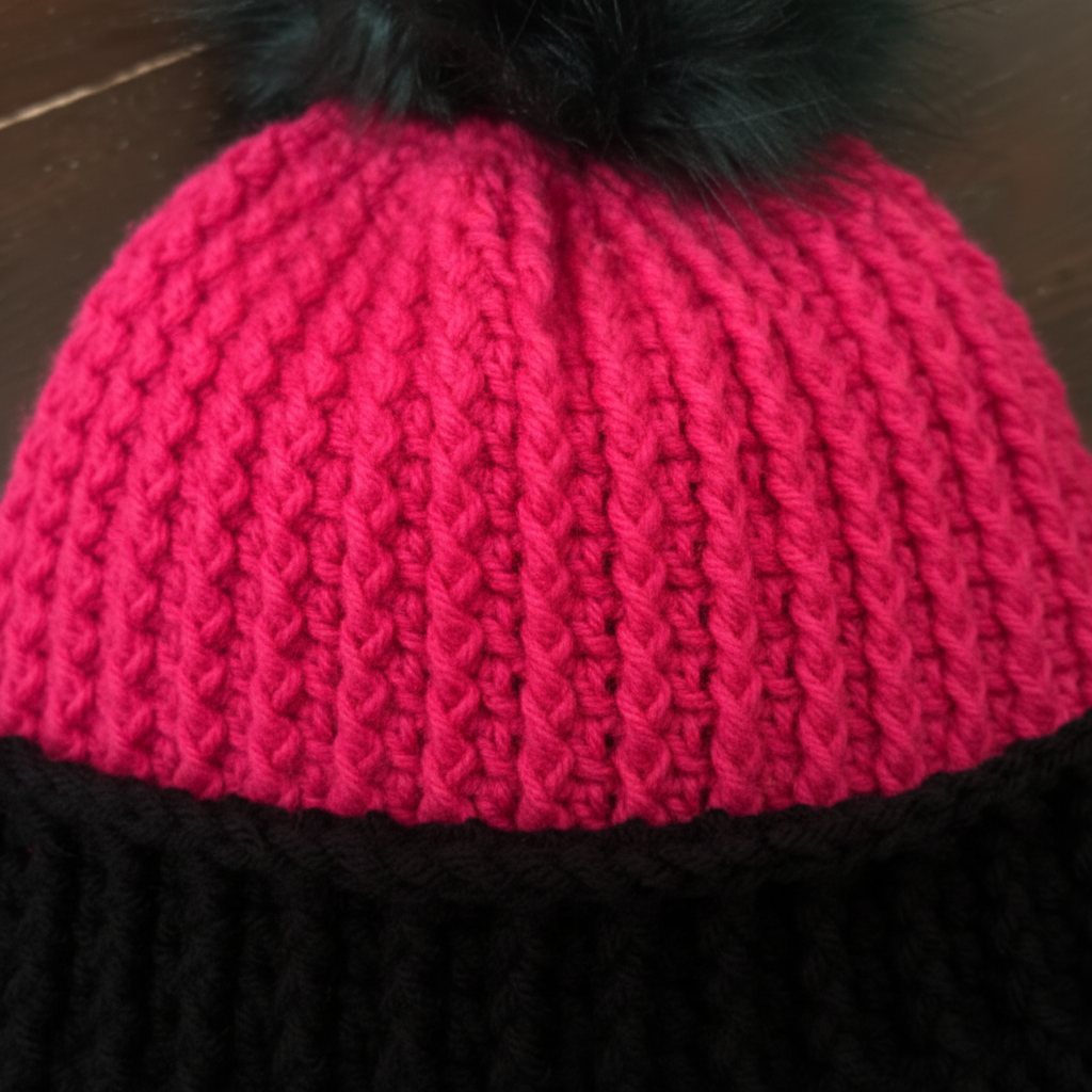 Pink crochet beanie with a black pom-pom on a wooden surface