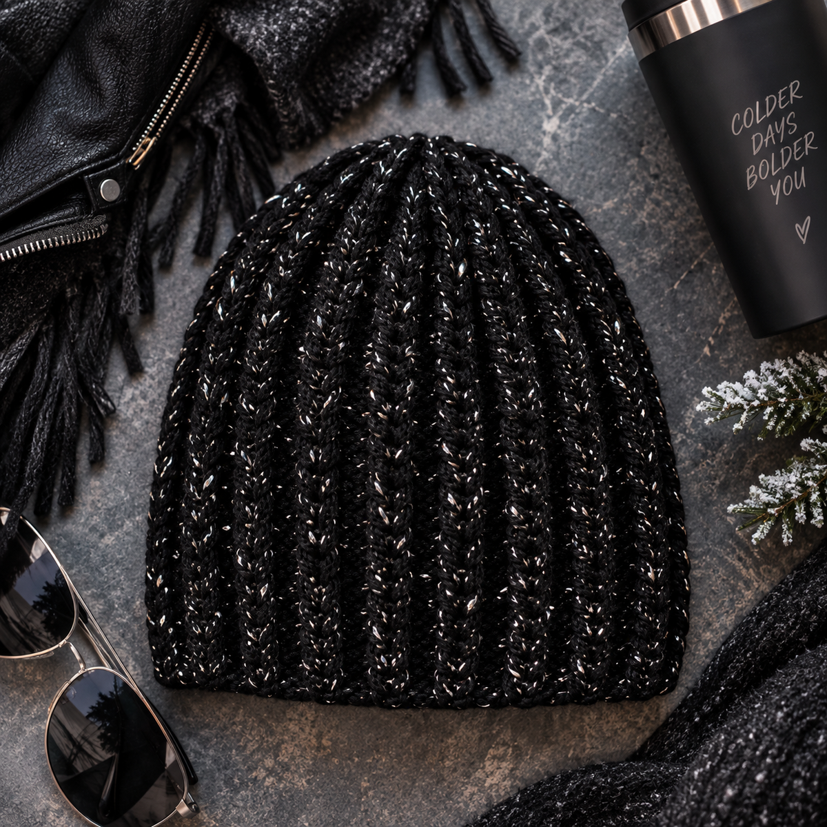 A black ribbed crochet beanie with subtle white flecks, styled in a flat lay on a textured gray surface with sunglasses, a black leather jacket, a fringed scarf, a black insulated tumbler, and small evergreen branches.