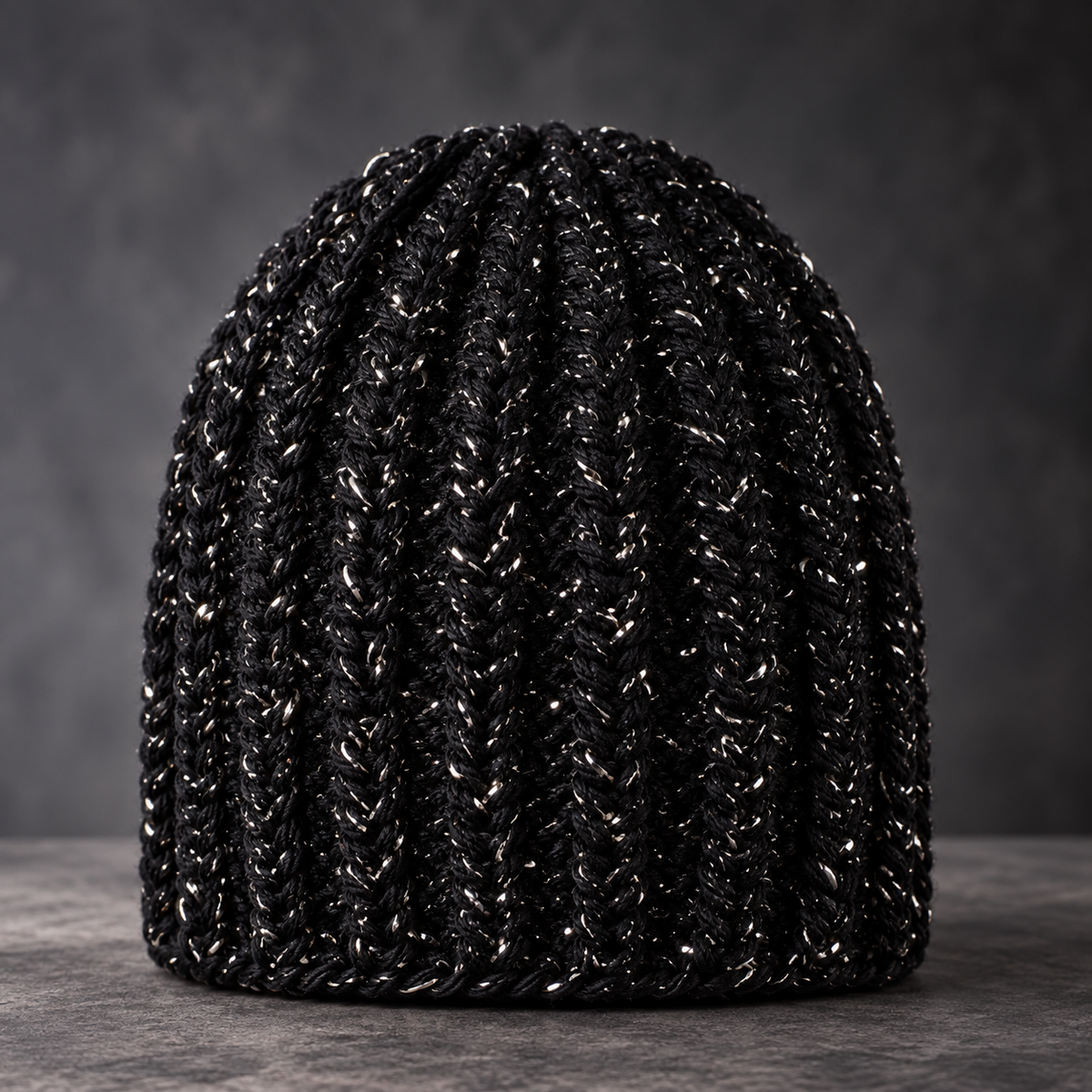 A black ribbed crochet beanie hat with subtle silver reflective flecks throughout, displayed on a dark gray textured surface against a dark gray background.
