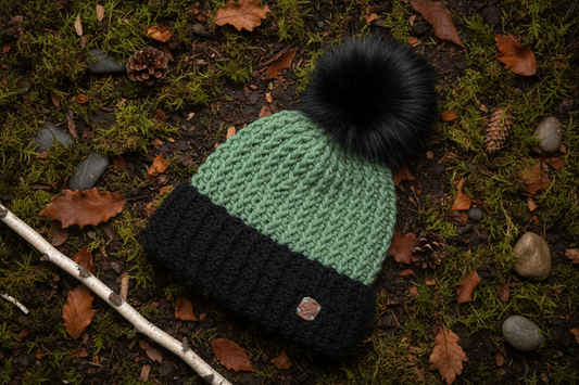A chunky crochet beanie hat with a black pom-pom, featuring a light green upper section and a black lower cuff. It is photographed from above on a forest floor background covered in moss, leaves, and pinecones.