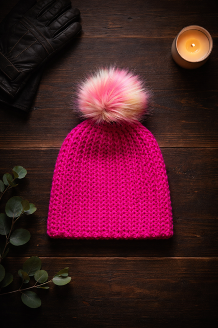Ignite crochet beanie in bold hot pink ribbed yarn with blush faux fur pom styled in warm flat lay composition on wooden surface for winter wear