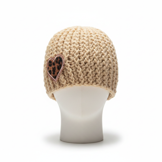 A beige chunky crochet beanie hat displayed on a white mannequin head. The hat features a textured v shape stitch pattern and a small heart-shaped patch on the side with a leopard print design and a rose gold border.