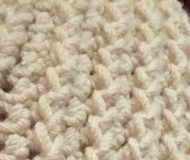 A close-up of a chunky, textured crochet stitch in a warm cream or off-white color.