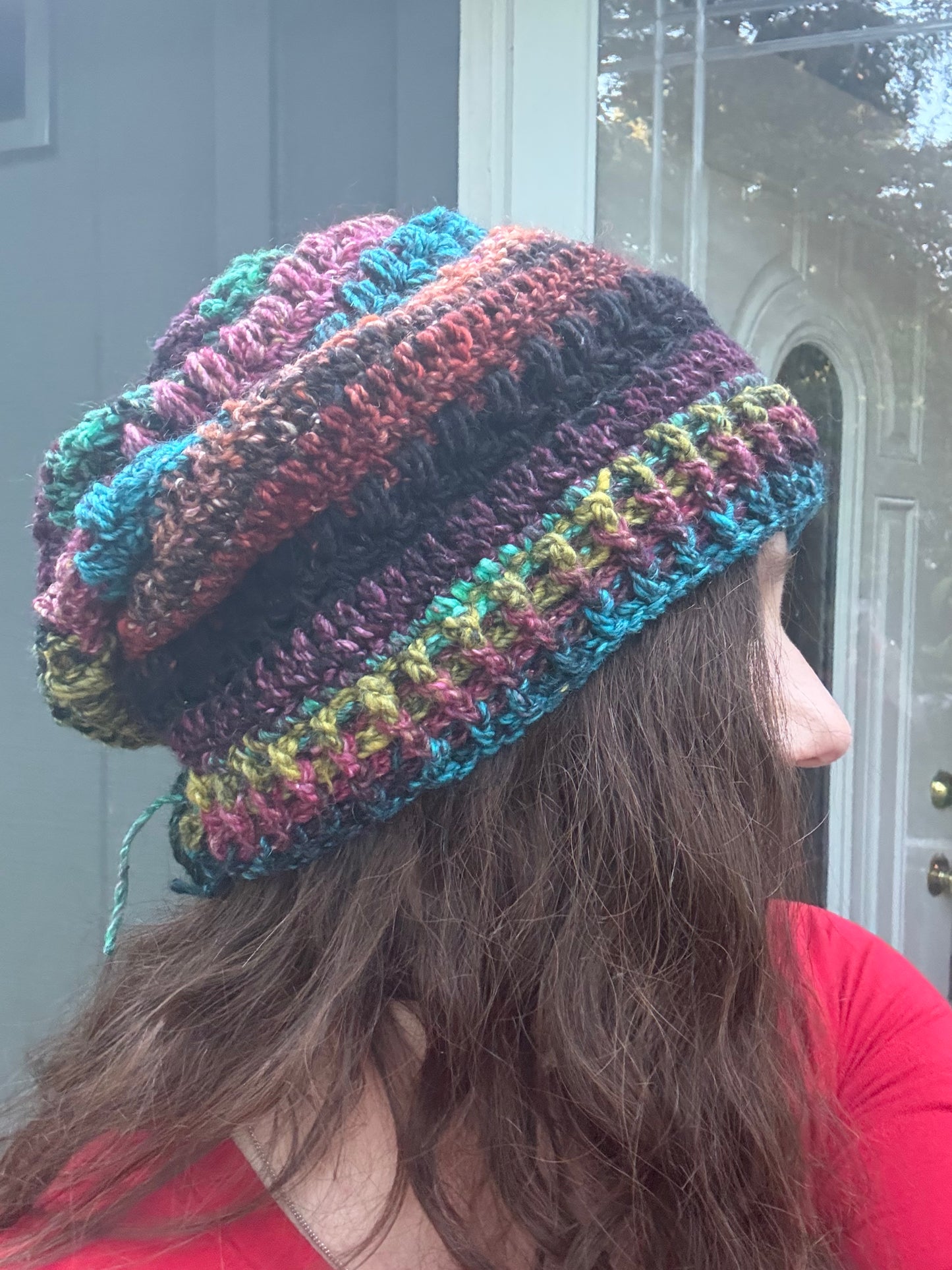 A person shown from the side wearing a chunky, multicolored crocheted slouchy beanie hat. The hat features stripes of teal, pink, purple, black, and olive green yarn.