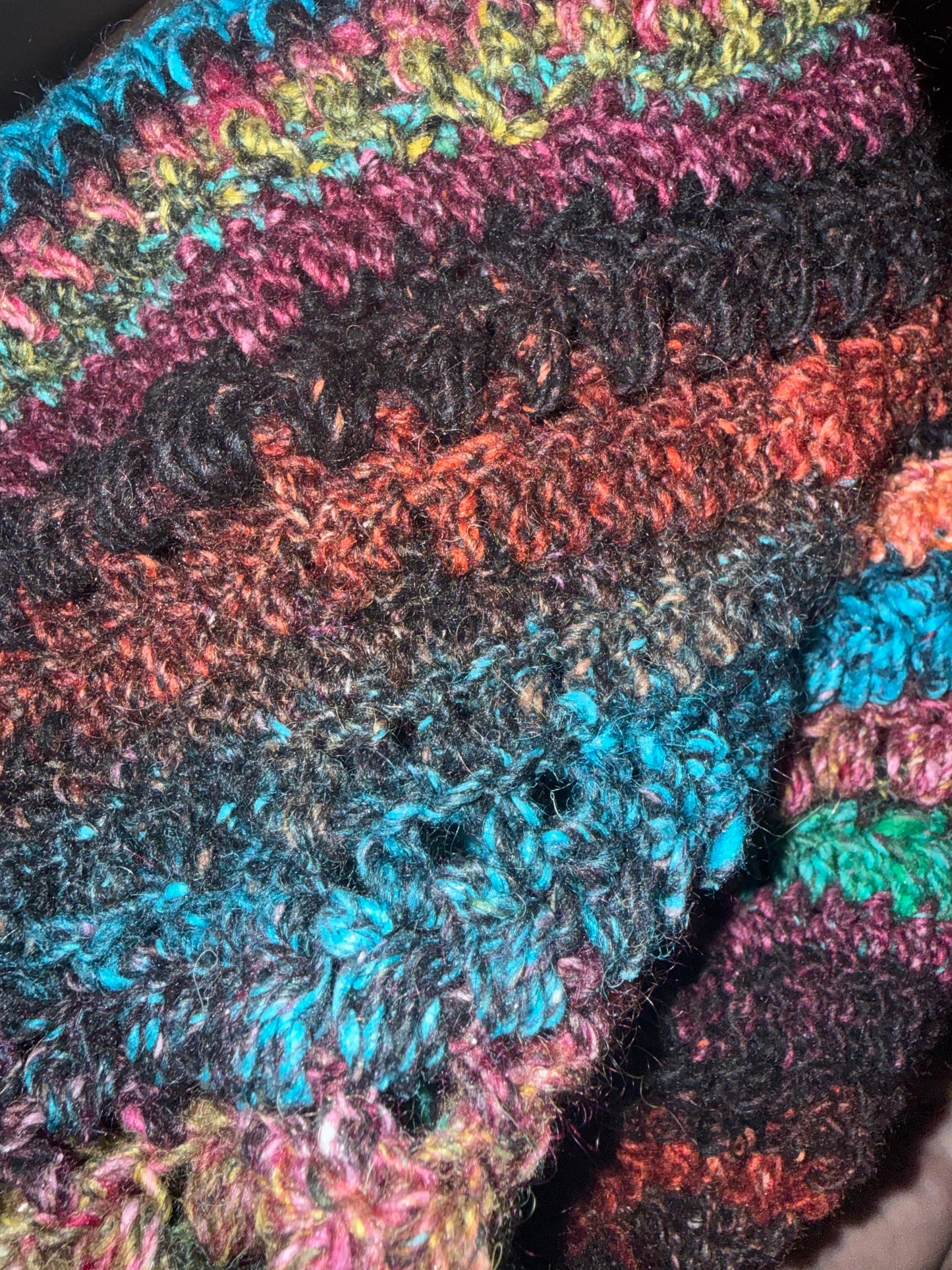 Close-up of a handmade, chunky crocheted textile featuring horizontal stripes of black, rust orange, teal blue, pink, and green yarn.