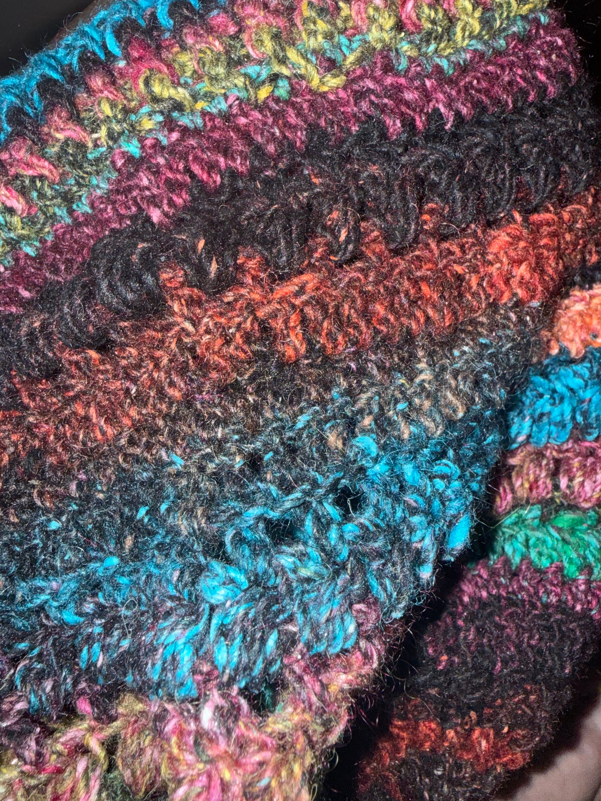 Close-up of a handmade, chunky crocheted textile featuring horizontal stripes of black, rust orange, teal blue, pink, and green yarn.